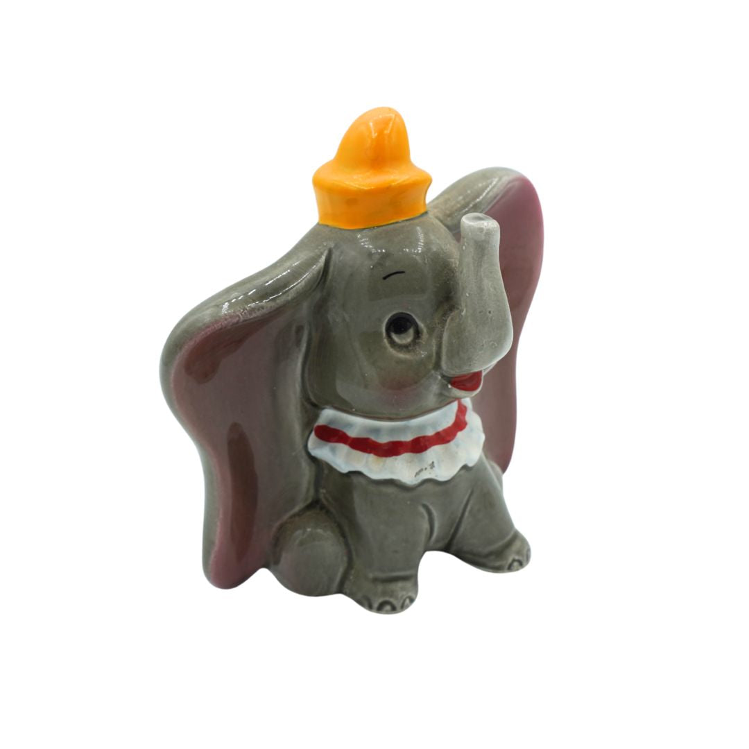 A small figurine of Dumbo the elephant wearing an orange hat and a white collar with red trim, set against a plain white background.