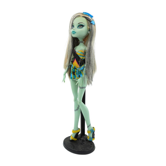 Monster High doll with long silver-blonde hair, blue eyes, and a green skin tone, wearing a black, blue, and yellow patterned outfit with yellow heels, displayed on a black stand against a white background.