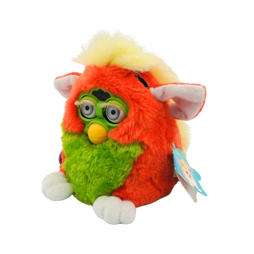An orange and green Furby with yellow hair and blue eyes, photographed against a white background