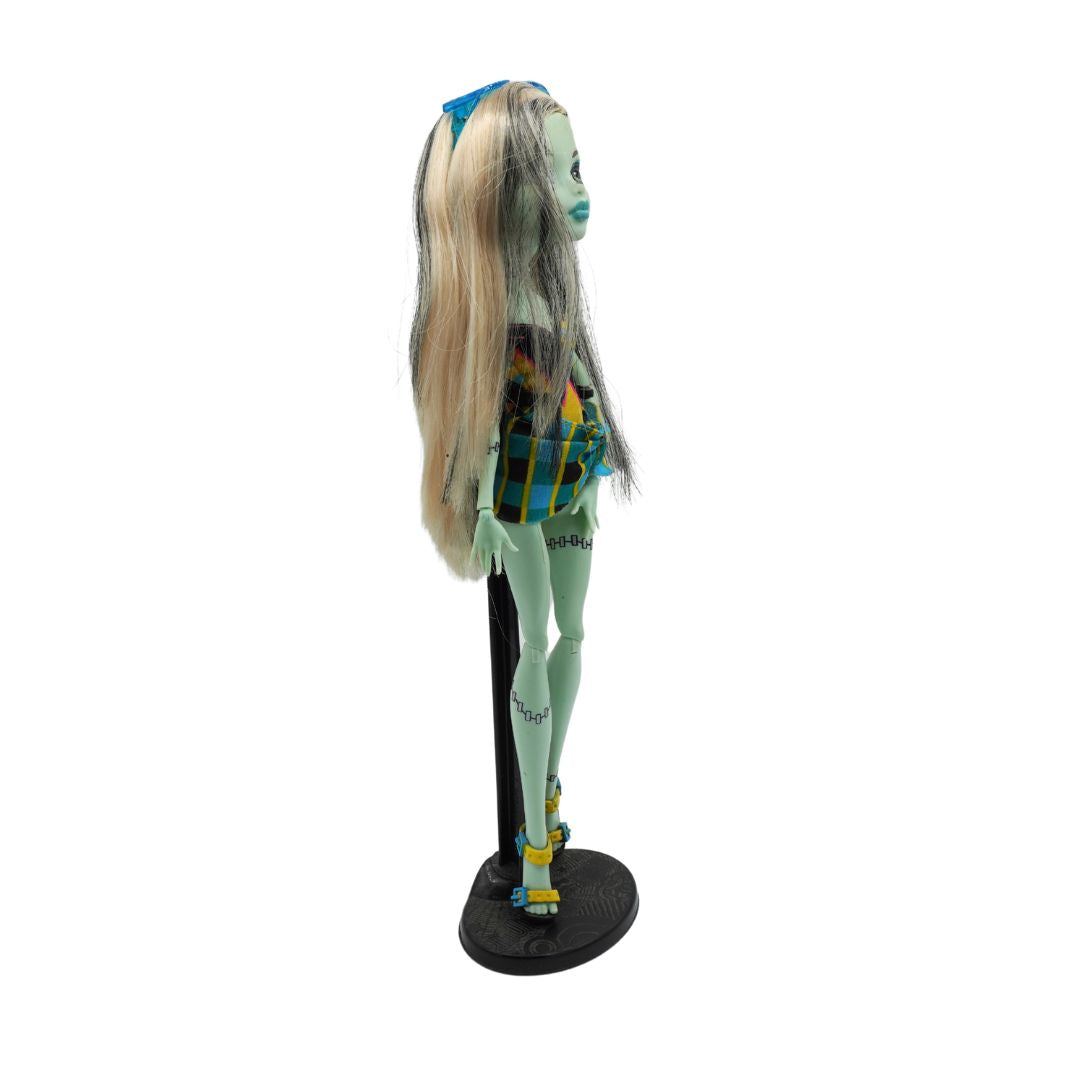 A Monster High doll with long blonde and gray hair, wearing a plaid top and light green tights, standing on a black display stand against a white background.