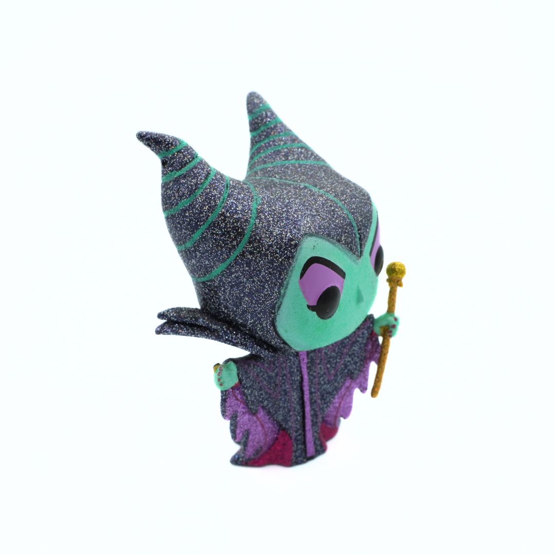 A chibi-style collectible figurine of Maleficent, featuring a glittery dark gray and purple outfit with teal accents, a green face, and a gold staff, set against a plain white background.