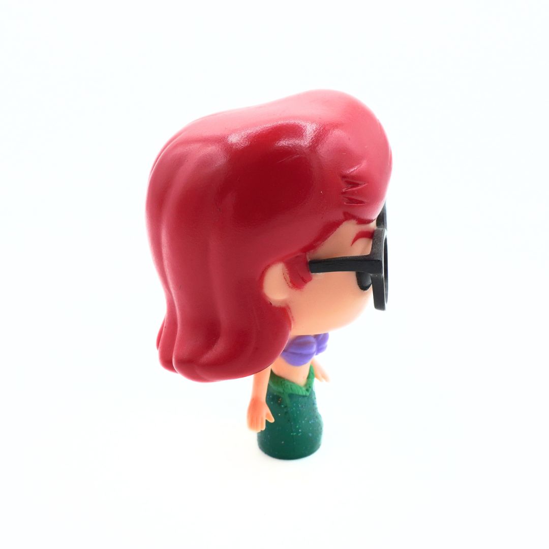 A stylized vinyl figure of a woman with short red hair, black sunglasses, a purple top, and a green skirt, shown from a side-back angle on a white background.