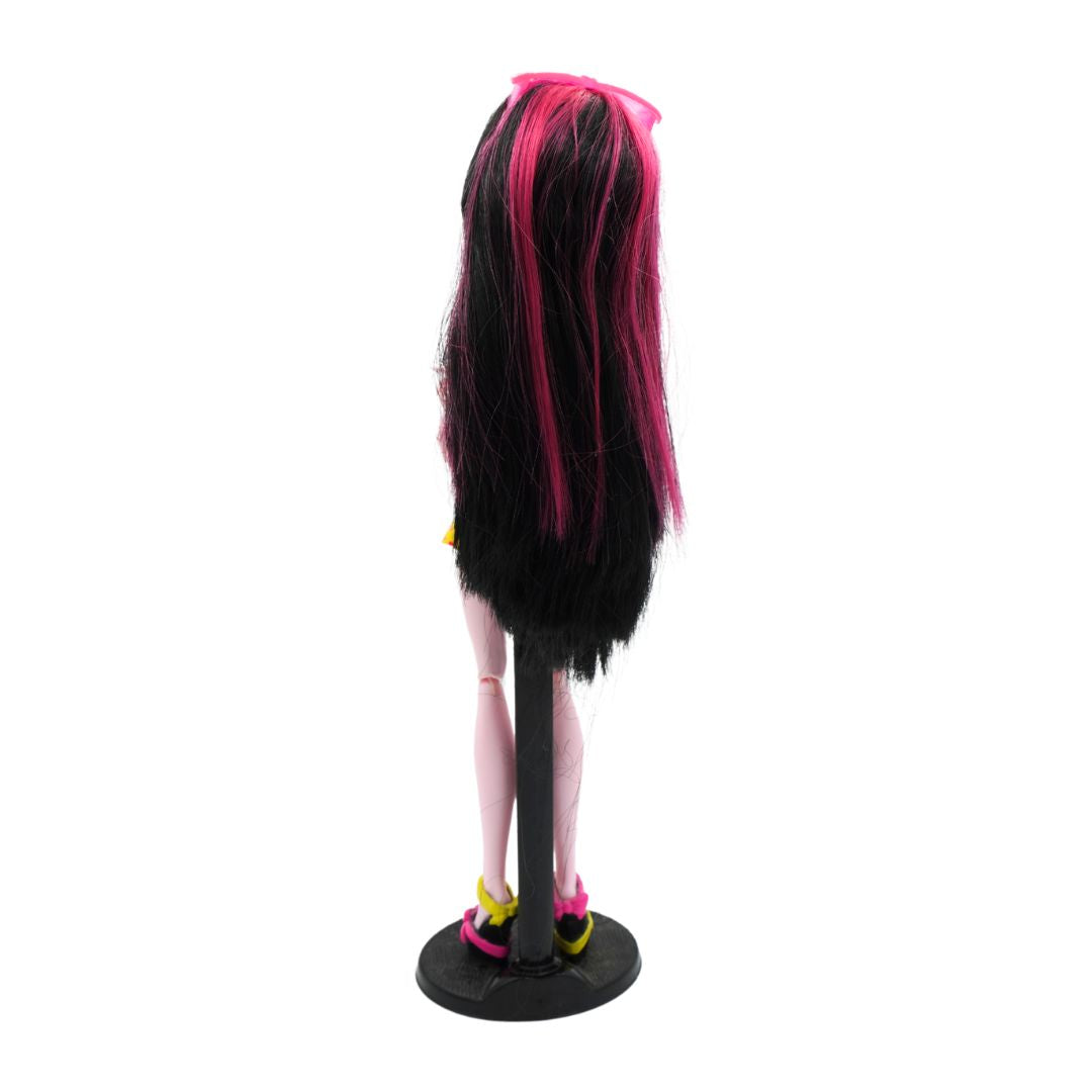 Back on photo of a 2010 Monster High Draculaura from the Gloom Beach range with black and pink hair, photographed on a black stand on a white background.