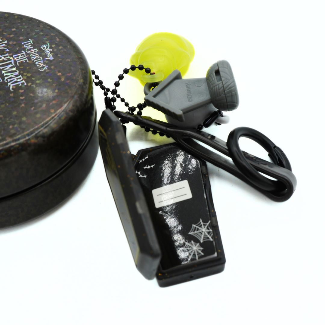 A Disney The Nightmare Before Christmas themed whistle keychain set, featuring a black metal tin with white text, a black plastic whistle with spiderweb graphics, a gray whistle cover, a black ball chain, and a translucent neon yellow earplugs accessory, all on a white background.