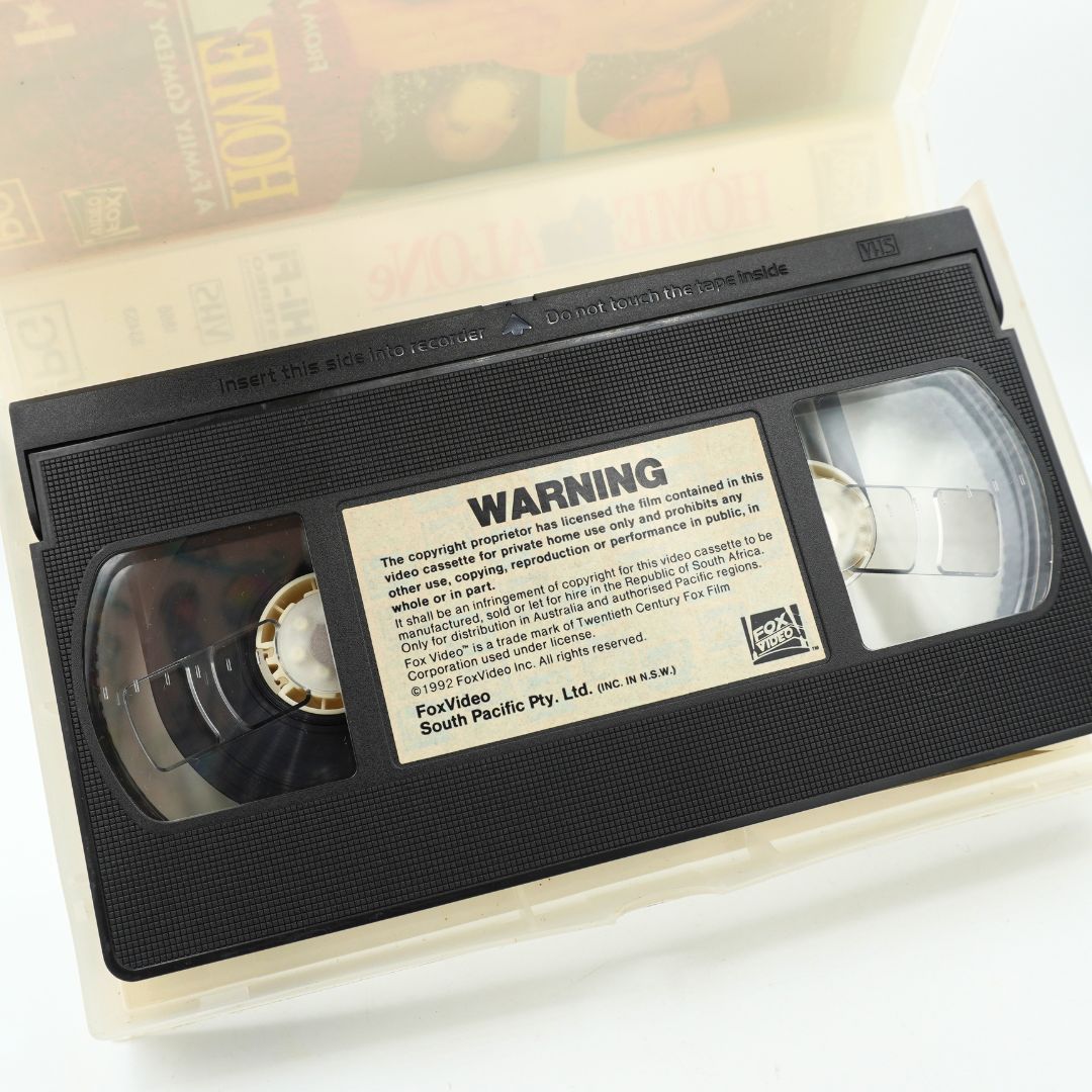 A vintage VHS video tape of the movie 'Home Alone' resting in its translucent plastic case, with the warning label clearly visible.