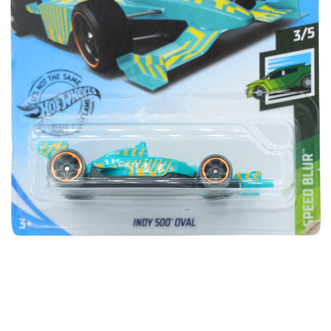A Hot Wheels die-cast toy race car in its original blister packaging, featuring a turquoise and yellow livery with 'INDY 500 OVAL' printed below on a white background.