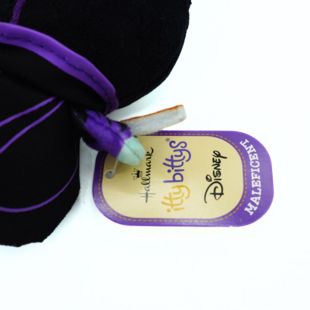 Close-up of a Disney Bitty's Maleficent plush toy with a black and purple fabric body, showing the attached Hallmark Disney Bitty's Maleficent hangtag on a white background.
