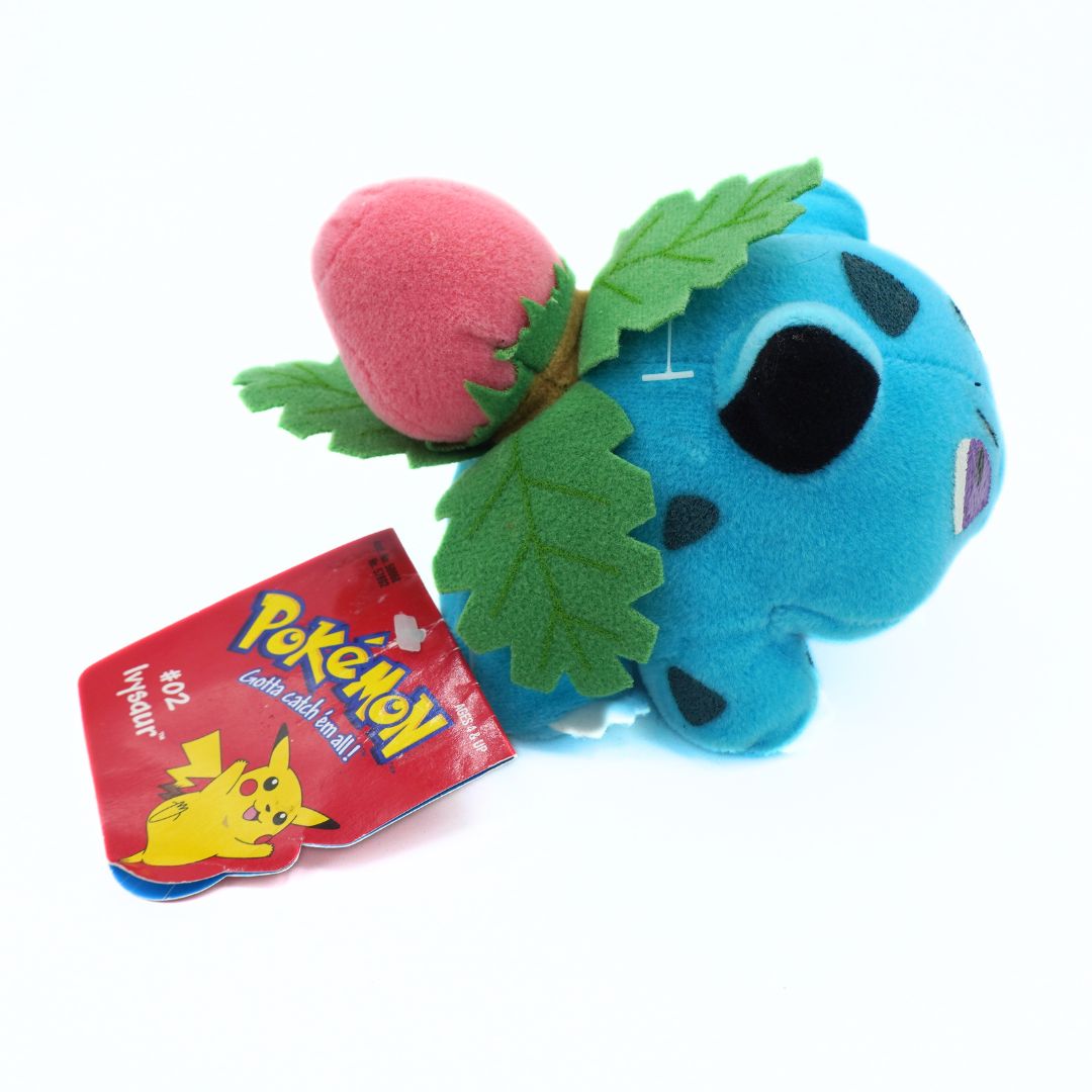 Blue plush toy with pink heart and green leaves next to a Pokémon trading card on a white background