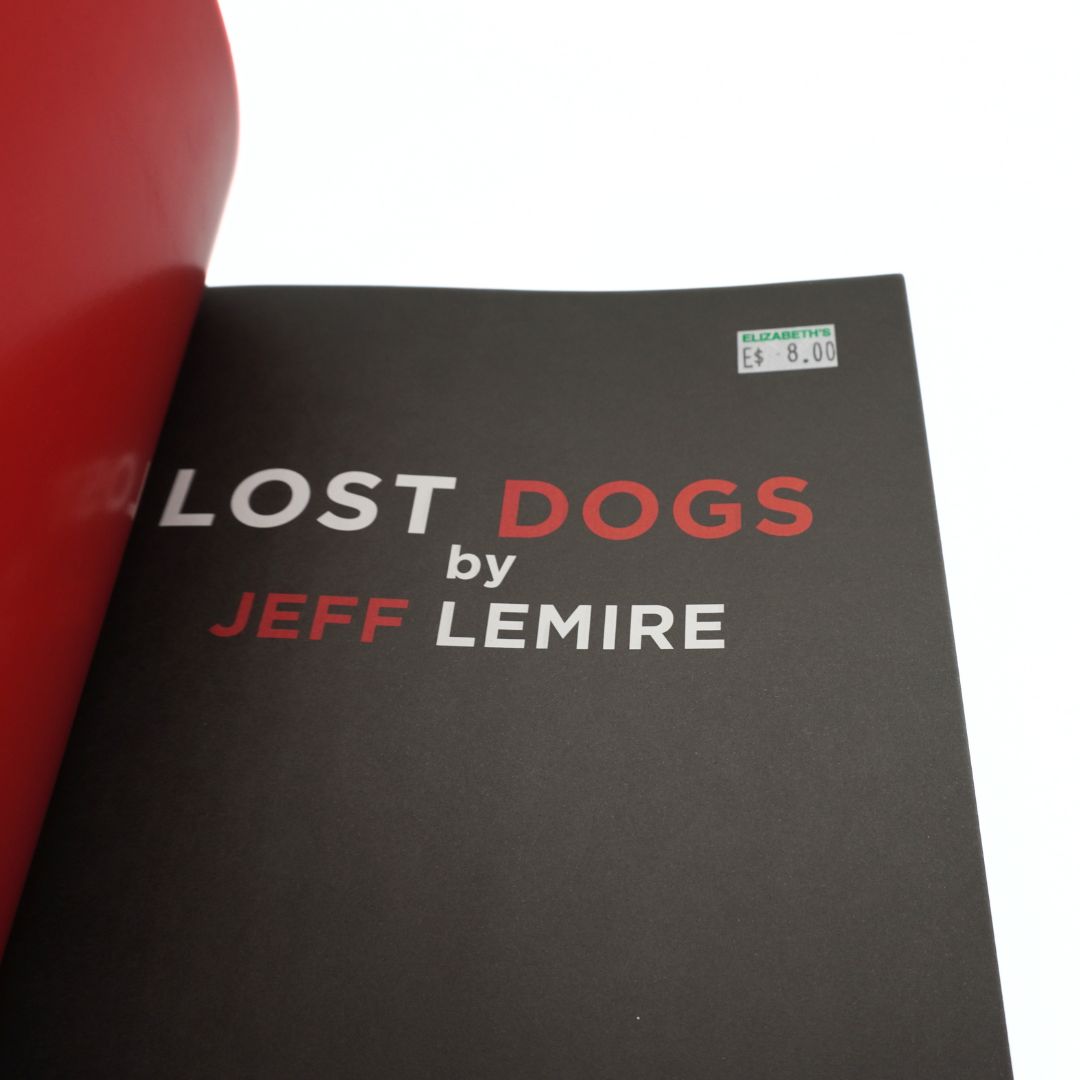 A close-up of a book cover titled "Lost Dogs by Jeff Lemire" on a white background, with part of a red object visible along the left edge.