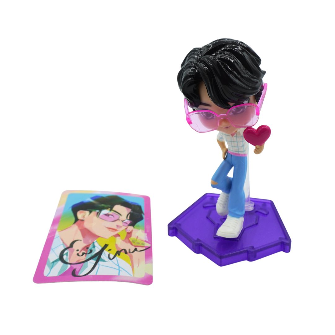 A small stylized figurine of a person with black hair, pink-tinted glasses, a white top, blue pants, white shoes, and a pink heart, standing on a purple base. A matching illustrated card is placed beside it on a plain white background.