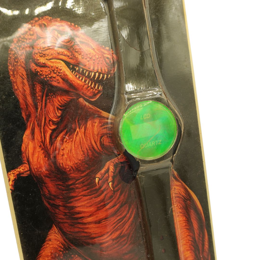 A wristwatch with a black strap and a bright green circular dial, shown attached to its retail backing card featuring a red dinosaur illustration.