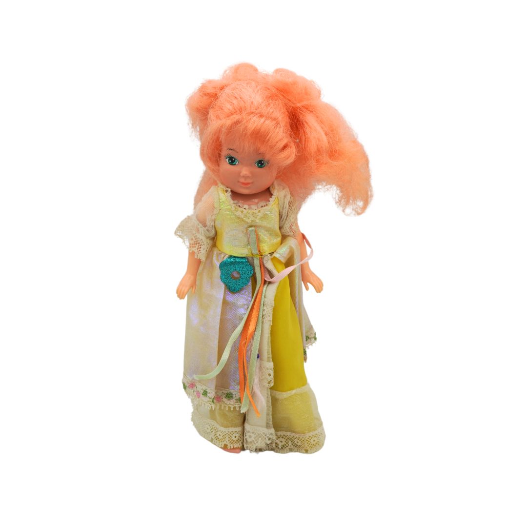 A collectible doll with orange hair styled in two high pigtails, wearing a yellow and white medieval-style dress with a turquoise flower accessory, set against a plain white background.