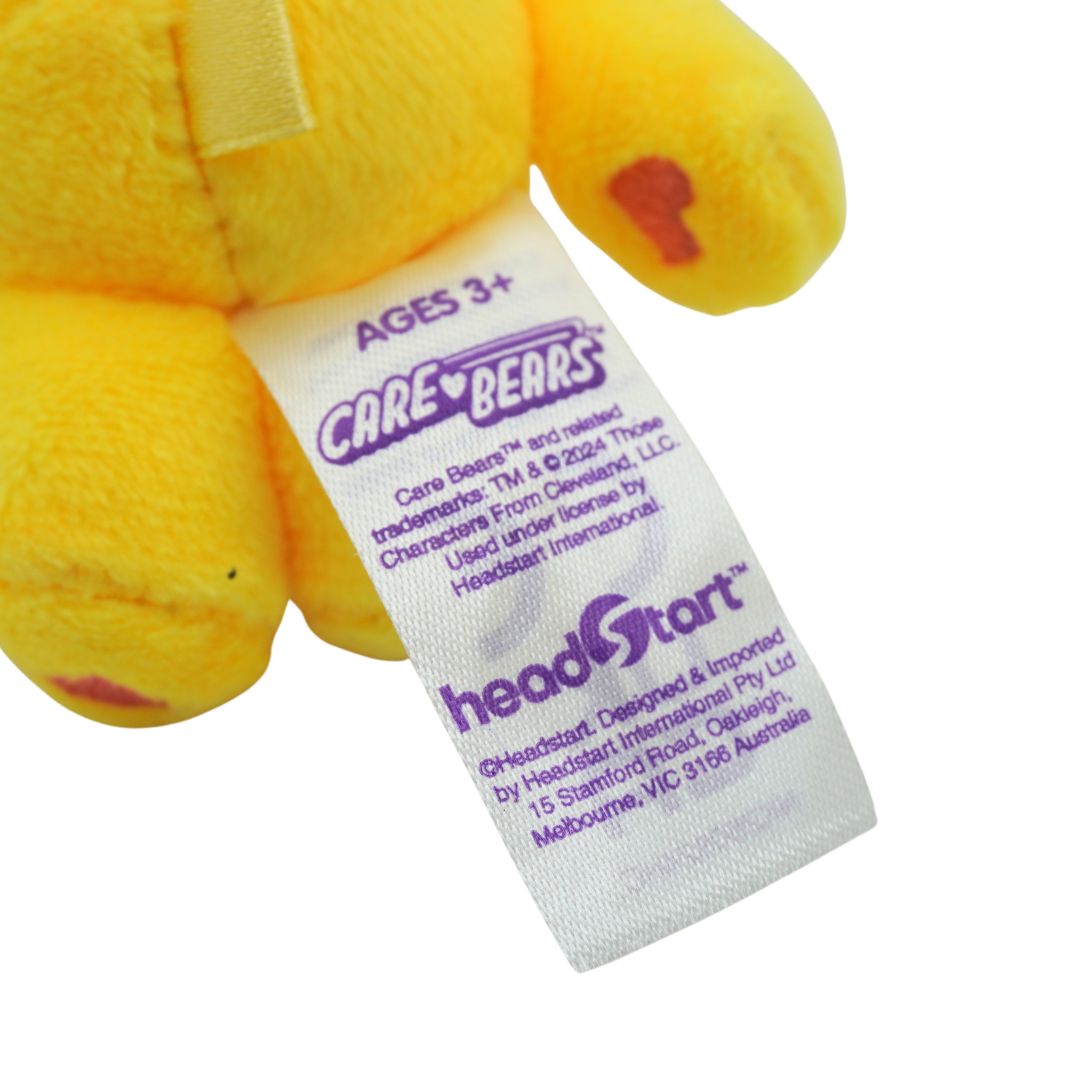 Close-up of a yellow plush toy with a sewn-on white Care Bears tag reading 'headStart' and 'Ages 3+'.