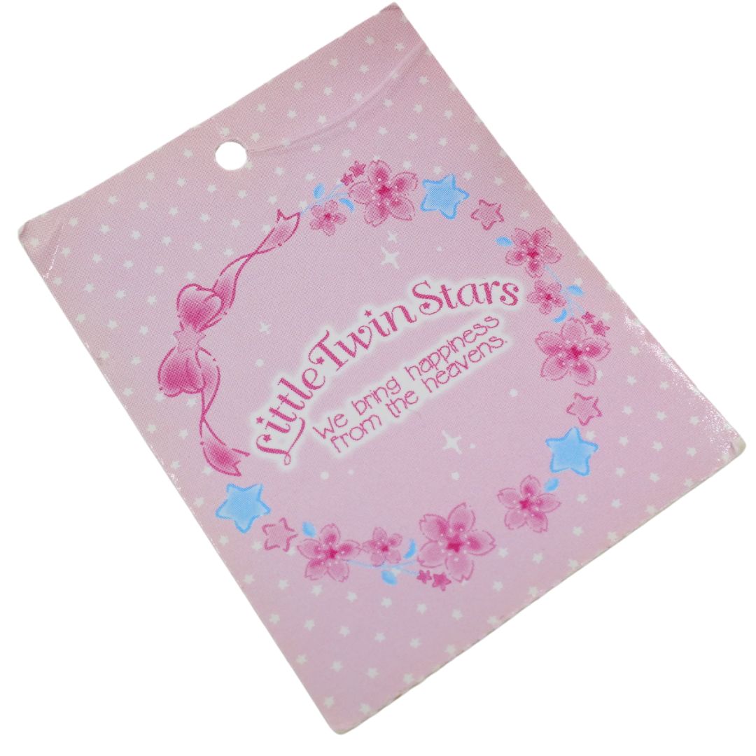 A square pink card or packaging insert with a hanging hole, printed with the text 'Little Twin Stars' and 'we bring happiness from the heavens' inside a floral wreath design with pink and blue stars on a white background.