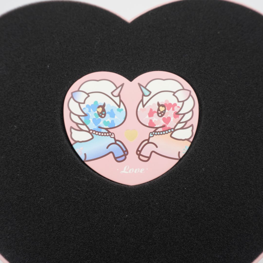 A heart-shaped pink coaster featuring two cute cartoon unicorns facing each other, displayed on a black heart-shaped background.
