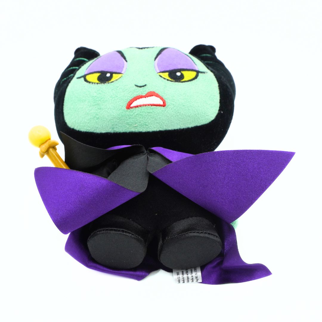 A plush doll of a female vampire character with a light green face, purple lips, black hair, a black dress, and a purple cape, holding a small gold wand, set against a plain white background.