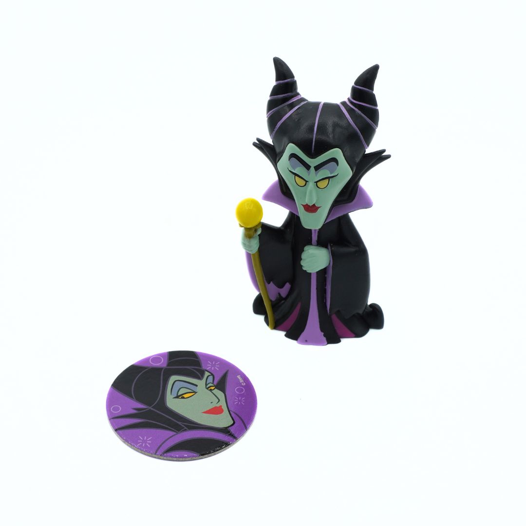 A small Maleficent figurine holding a yellow-tipped staff stands next to a round pin or sticker featuring a close-up of Maleficent's face and purple horned silhouette, on a plain white background.