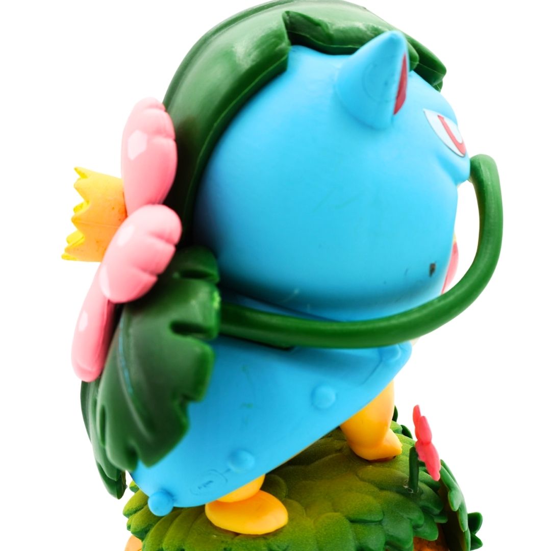 A close-up of a blue cartoon character figurine with green leafy hair, pink flowers, and yellow feet, shown against a plain white background.