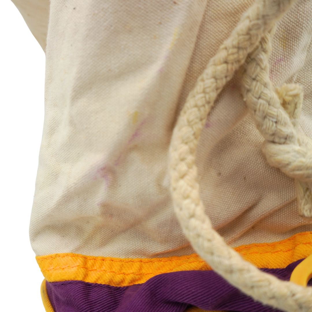 Close-up of a canvas drawstring bag with a cream-colored woven rope and a purple and orange fabric trim at the bottom, shown against a white background.