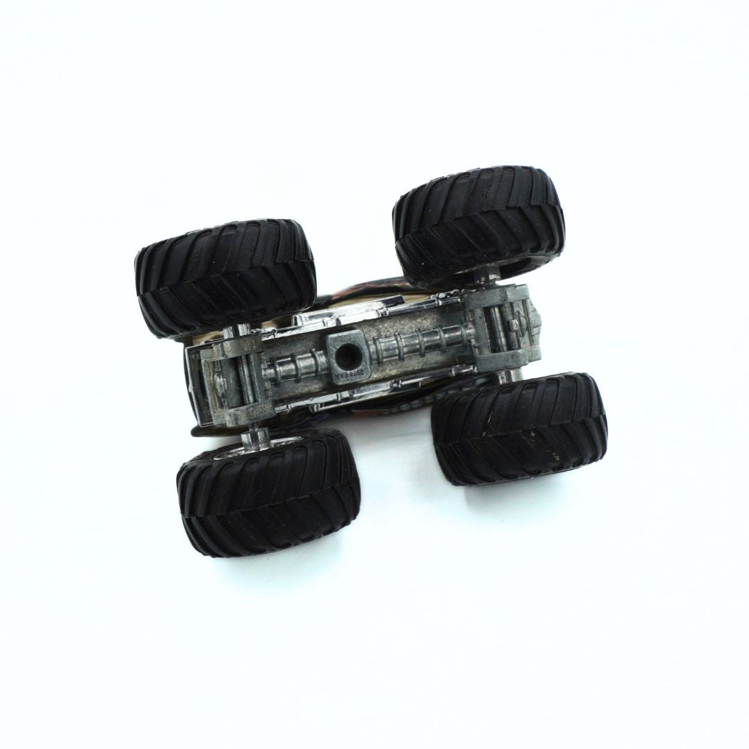 A miniature toy monster truck shown upside down, revealing its detailed gray chassis and four black off-road tires, set against a plain white background.