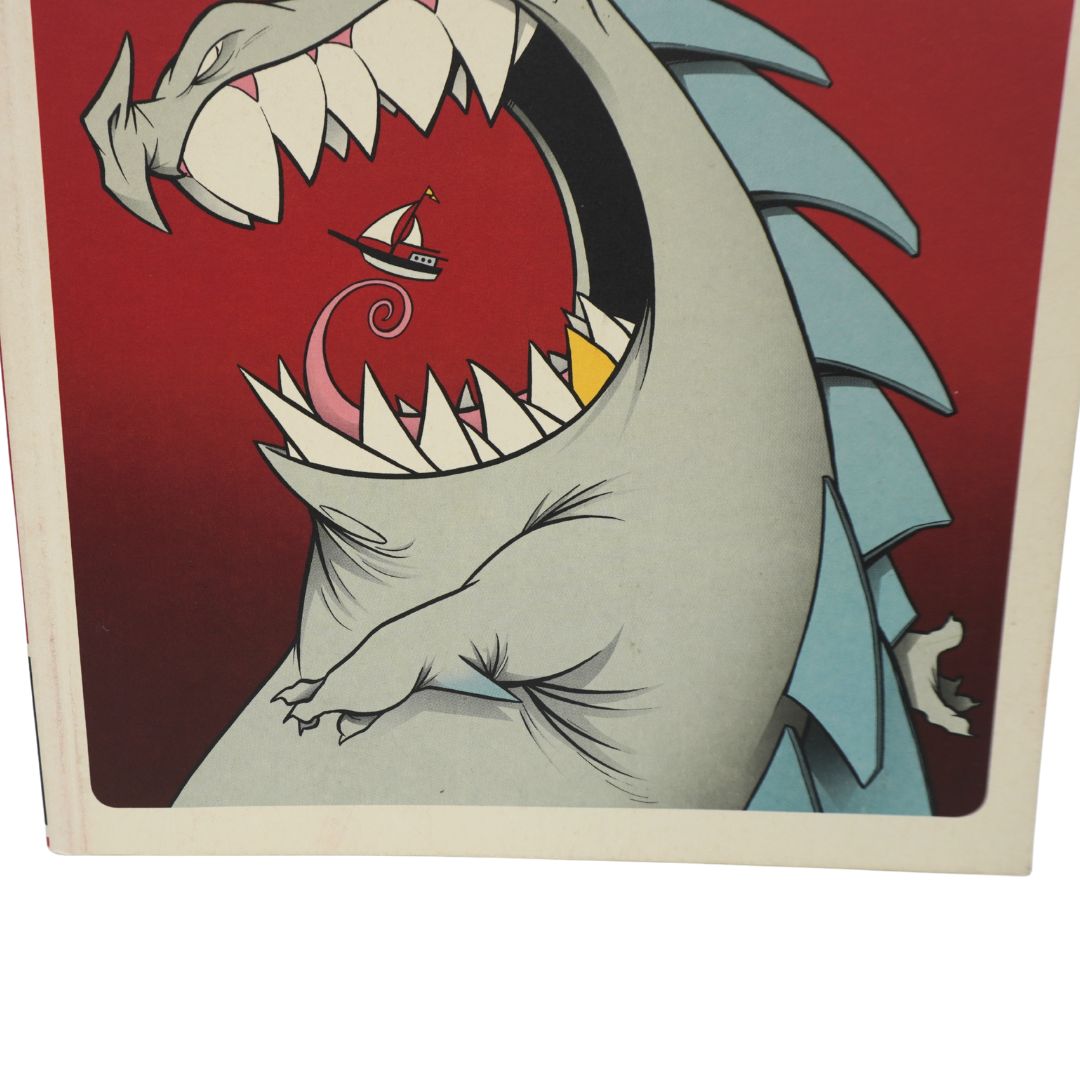 A close-up of a printed illustration showing a large gray monster or dragon with an open mouth, white teeth, and blue spikes against a red background, with a tiny boat inside the mouth.