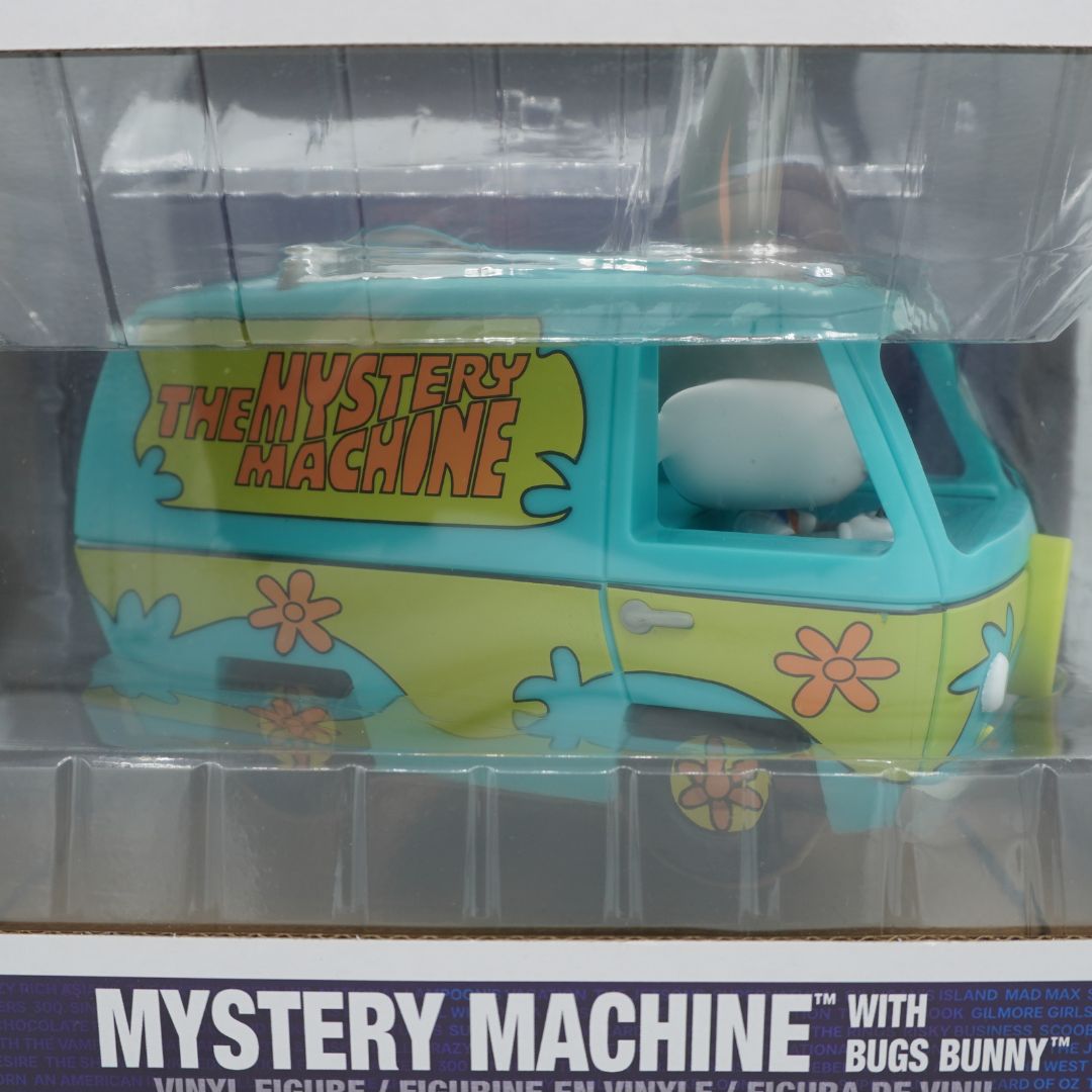 The Mystery Machine toy van from The Simpsons, packaged in a clear plastic blister pack with a cardboard backing. The van is light blue and lime green with pink flowers and 'THE MYSTERY MACHINE' text on the side, and a Bugs Bunny figure is visible inside.