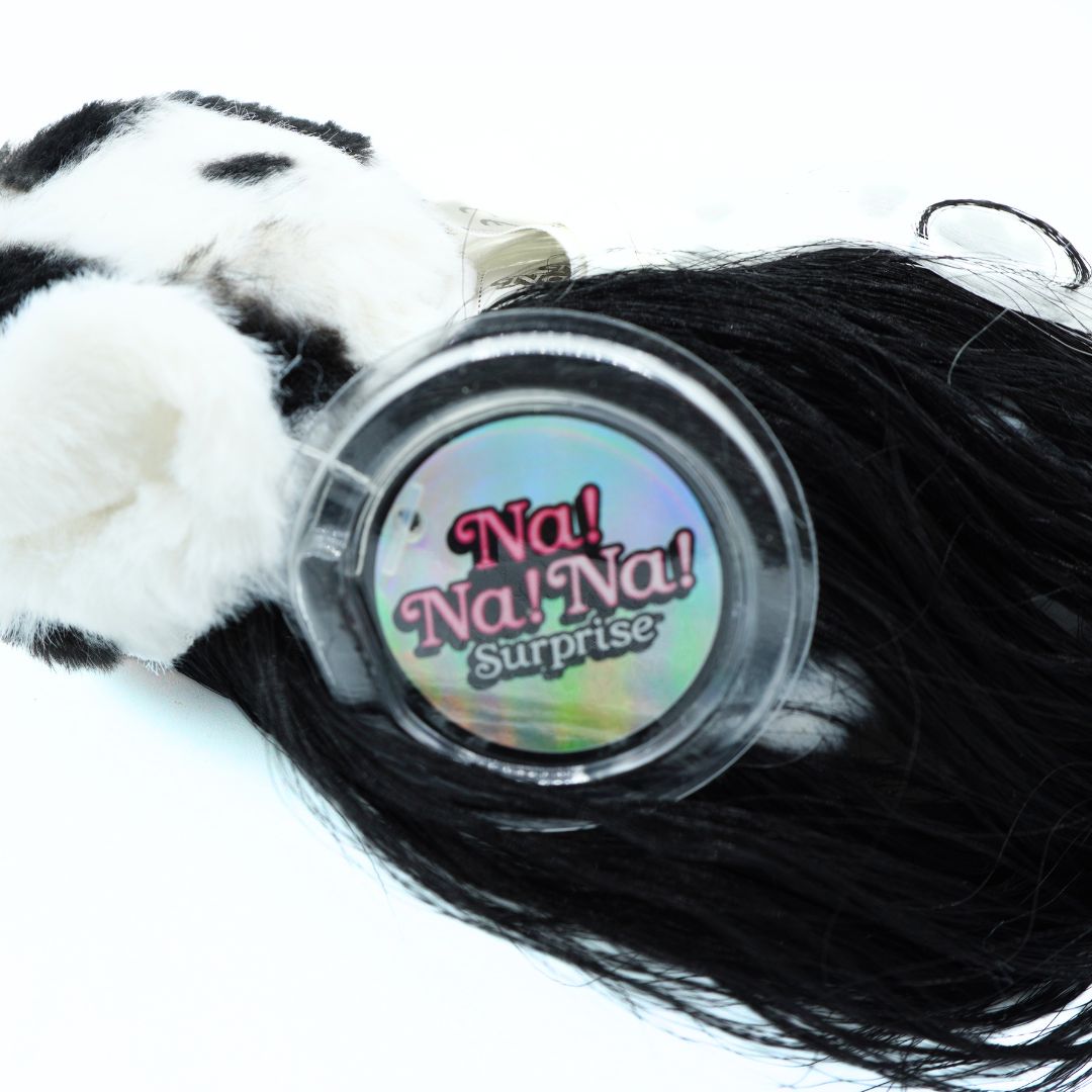 A close-up of a black faux fur or wig-like product wrapped in a clear plastic sleeve with a round label reading 'Na! Na! Na! Surprise'. The left side shows white and black fuzzy material, and the product is photographed on a plain white background.