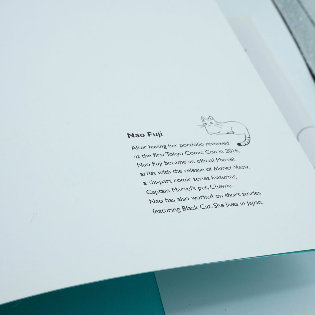 A close-up of an open book or booklet page with printed text about Nao Fuji and a small line drawing of a cat, photographed against a white interior with a teal page edge visible at the bottom.