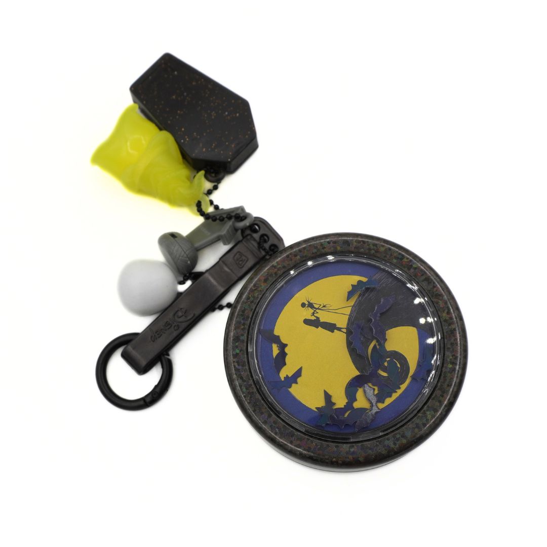 A Halloween-themed tin keychain featuring a round design with a dark blue bat silhouette against a yellow full moon, complete with a black textured hexagonal charm, a translucent neon yellow earplugs keyring, and a small white ball accent on a white background.