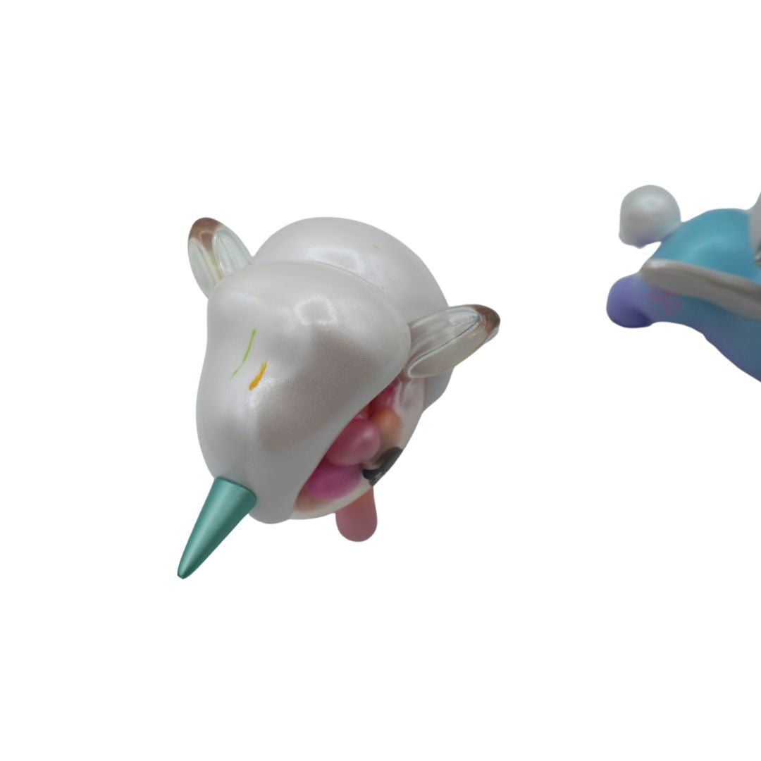 A white unicorn toy with a metallic teal horn and small embroidered yellow and green details on its forehead, shown against a plain white background. A second blue and purple plush toy is partially visible on the right edge.
