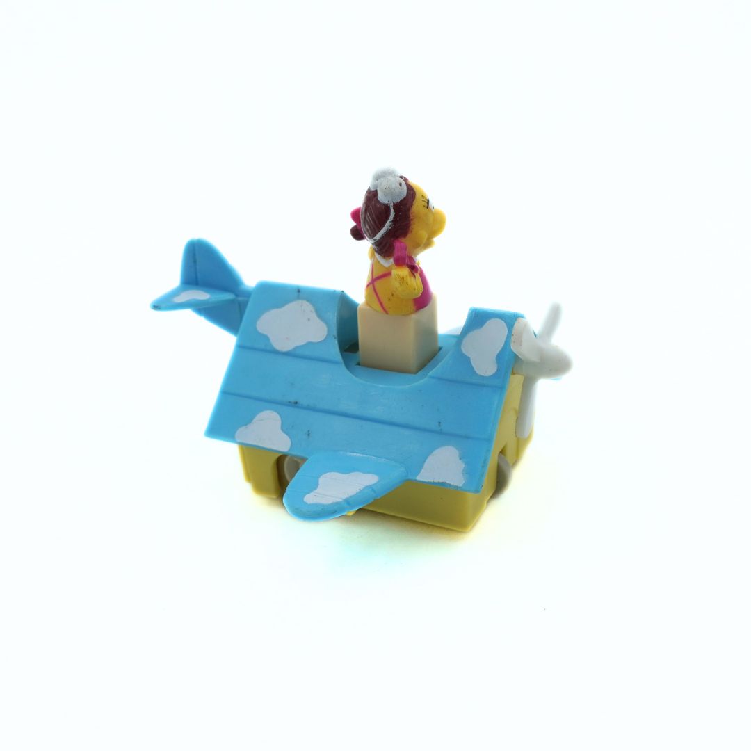 A small toy airplane-shaped container or playset with a yellow base, blue roof, white cloud decals, and a small yellow bird figure wearing a pink outfit and white hat standing in the cockpit, photographed on a white background.