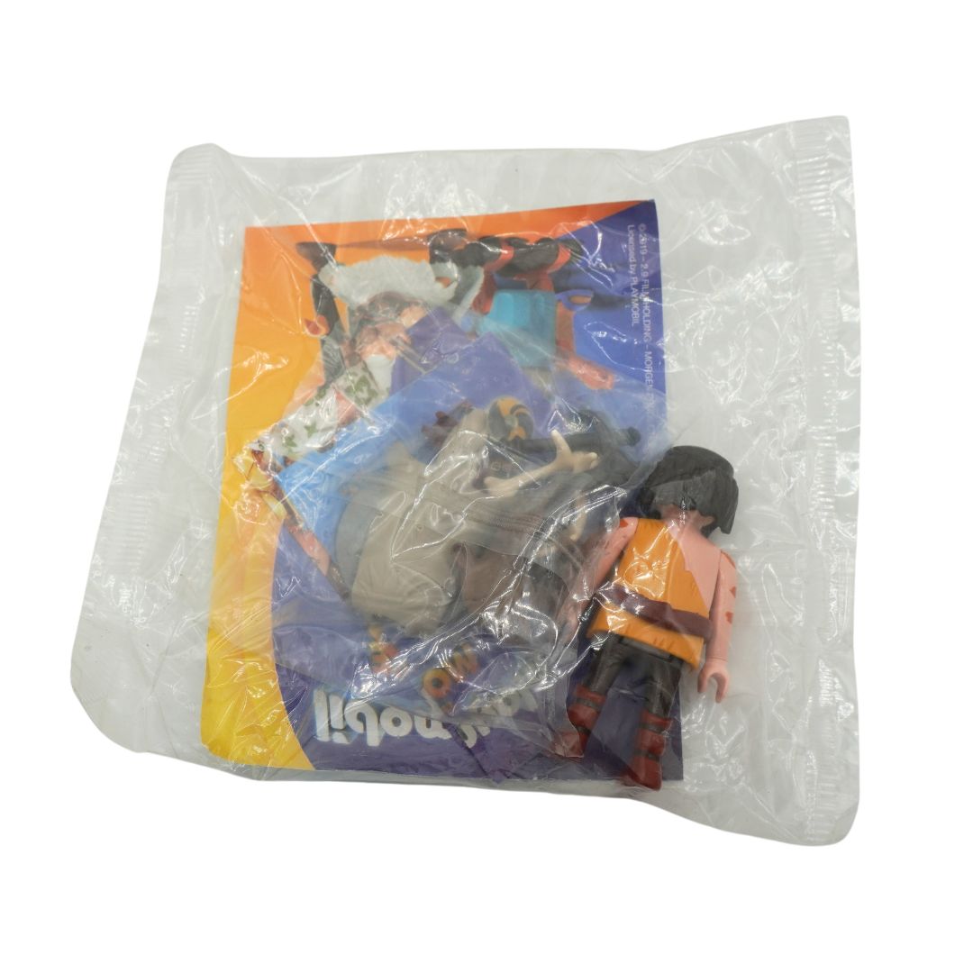 A small toy figure in an orange and black outfit standing next to its printed backing card, both sealed inside a clear plastic bag on a white background.