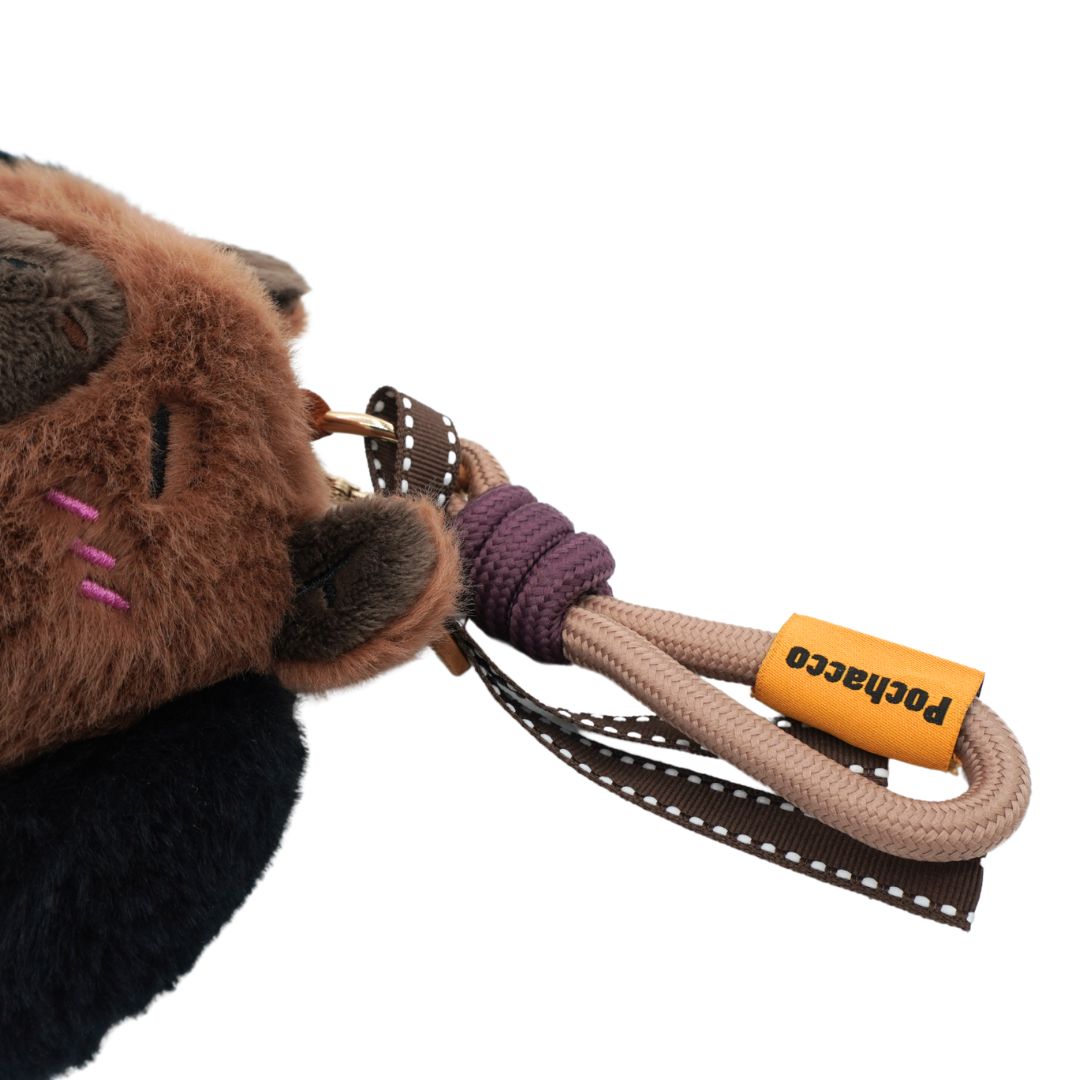 A close-up of a plush keychain or bag charm with a brown furry face, dark nose and ears, pink stitched whiskers, a beige rope loop, a purple wrapped section, and a small yellow tag reading Pochacco.