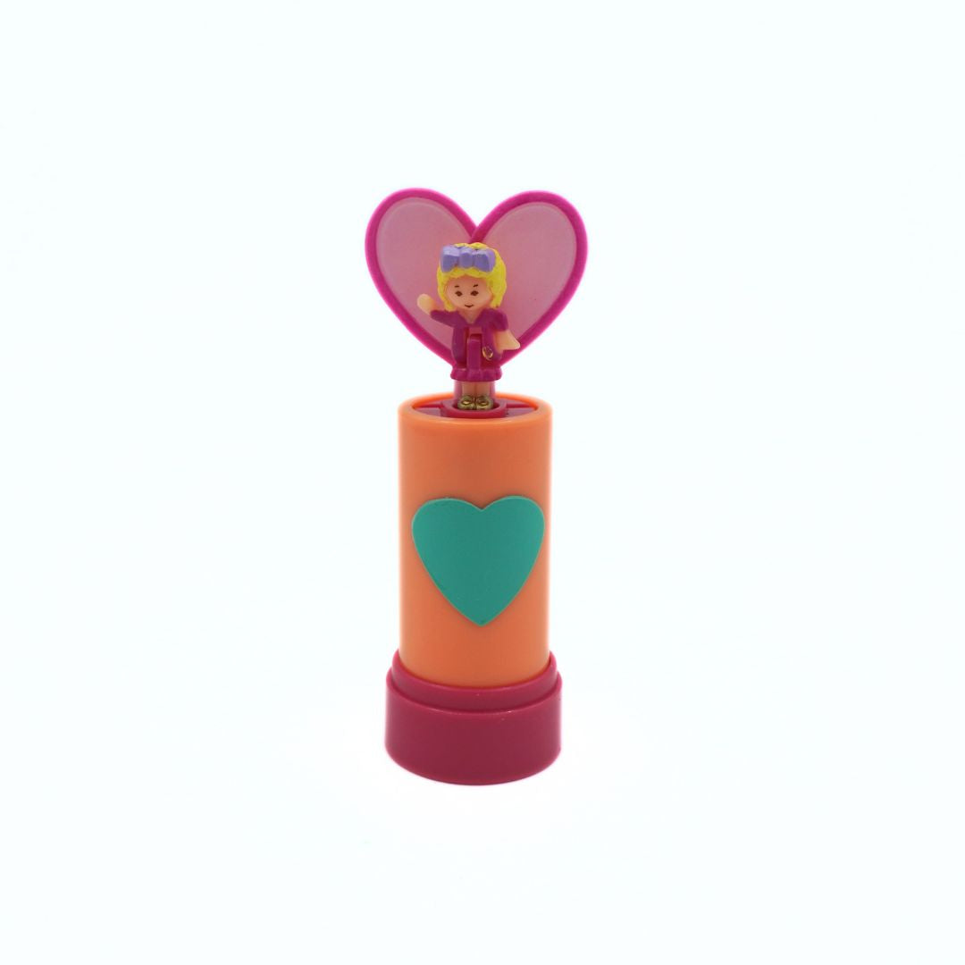 A small plastic toy wind-up jack-in-the-box with an orange body, pink base, teal heart on the front, and a pink heart-shaped opening showing a small blonde character.
