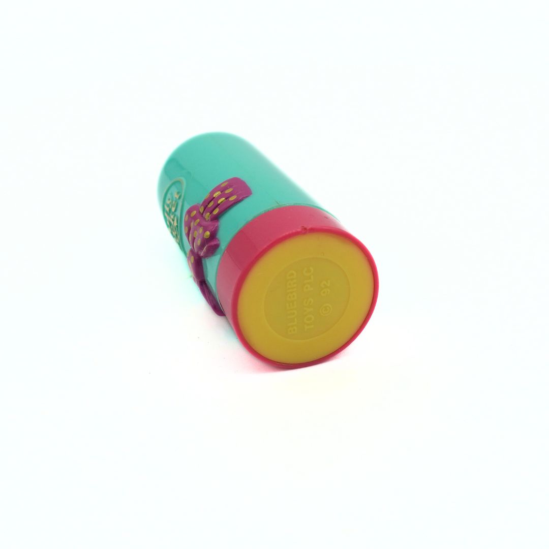 A colorful cylindrical cosmetic container, likely a lip balm tube, shown on a plain white background. The container features a teal-green body, a red rim, a yellow end cap, and a purple ribbon tied around the middle.