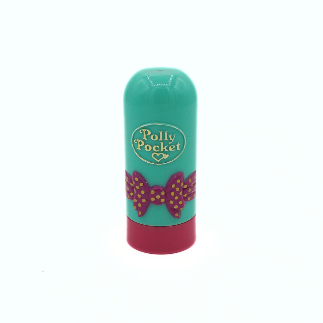 Polly Pocket lip balm stick with a turquoise green body, a pink bow design with gold polka dots, and a dark pink cap, shown on a white background.