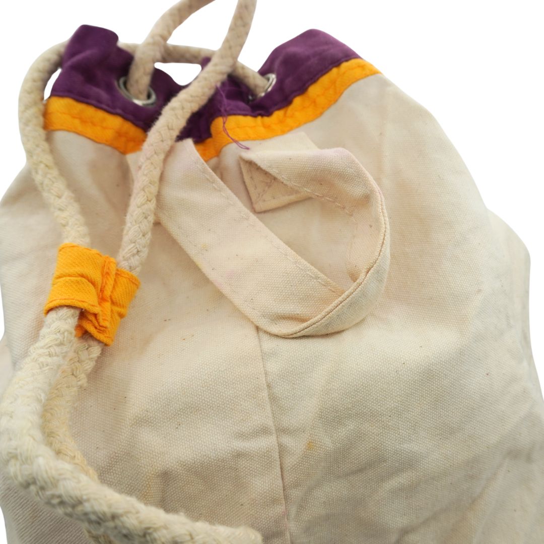A close-up of a canvas drawstring bag in natural beige fabric, featuring purple and orange trim at the top and thick braided rope handles.