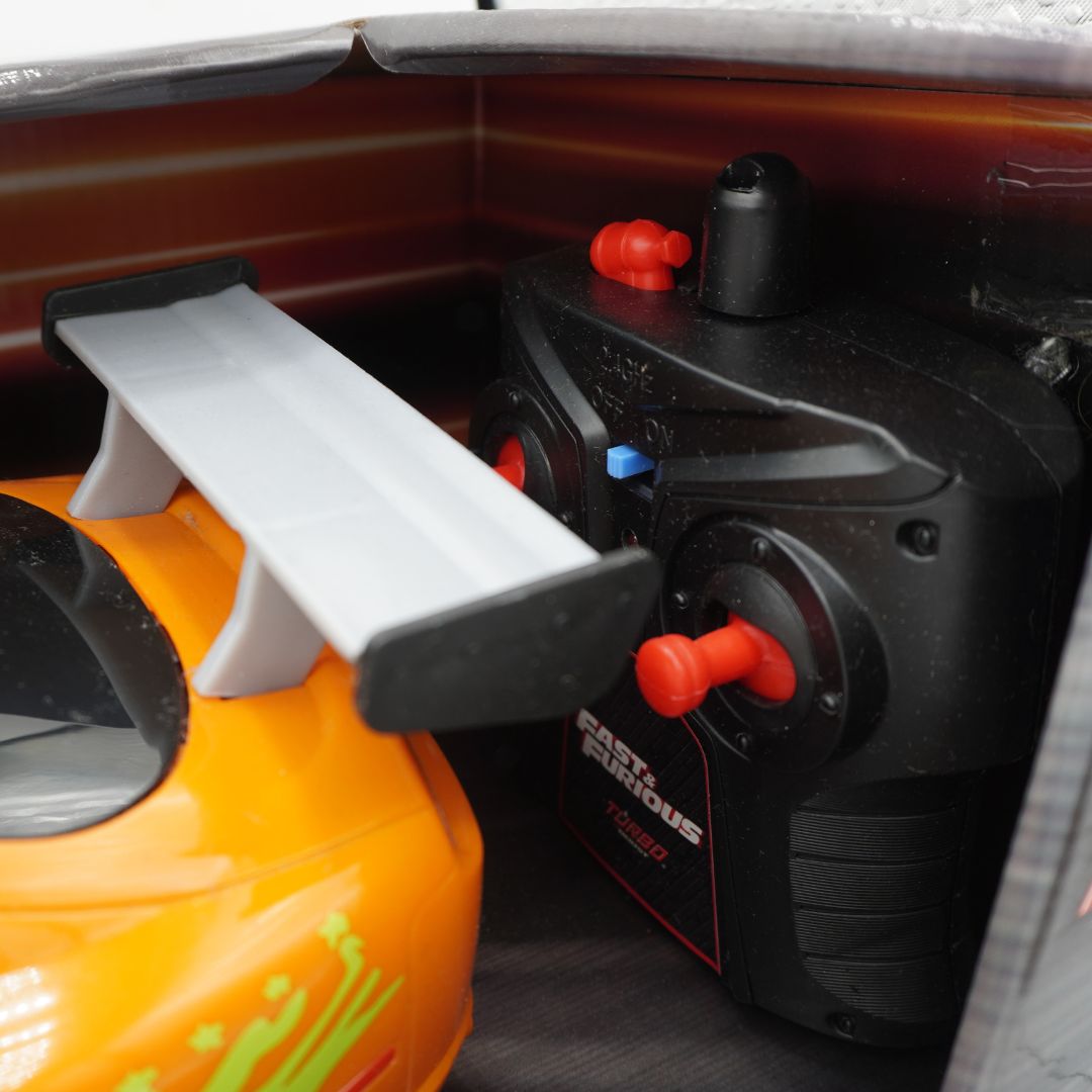 A close-up of a black remote control car transmitter with red joysticks and a white rear spoiler, placed next to an orange toy race car inside a storage compartment.