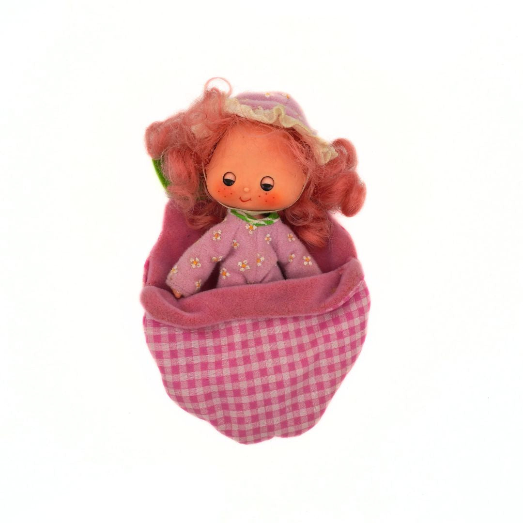Small doll with red hair and a pink dress inside a pink checkered bag on a white background