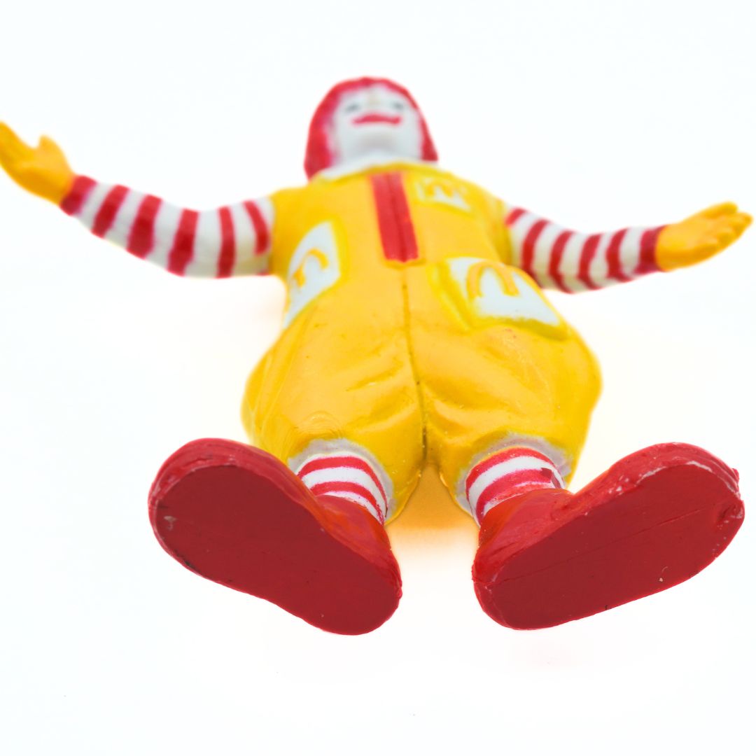 A small Ronald McDonald figurine posed lying on its back with arms outstretched, on a plain white background.