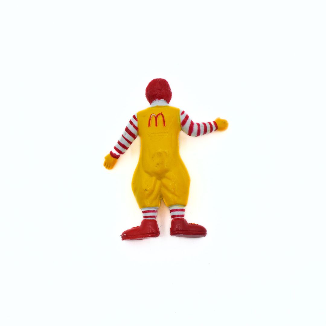 A Ronald McDonald toy figure shown from the back, wearing a yellow jumpsuit with the McDonald's golden arches logo on the back, red and white striped sleeves and socks, and red shoes, set against a plain white background.
