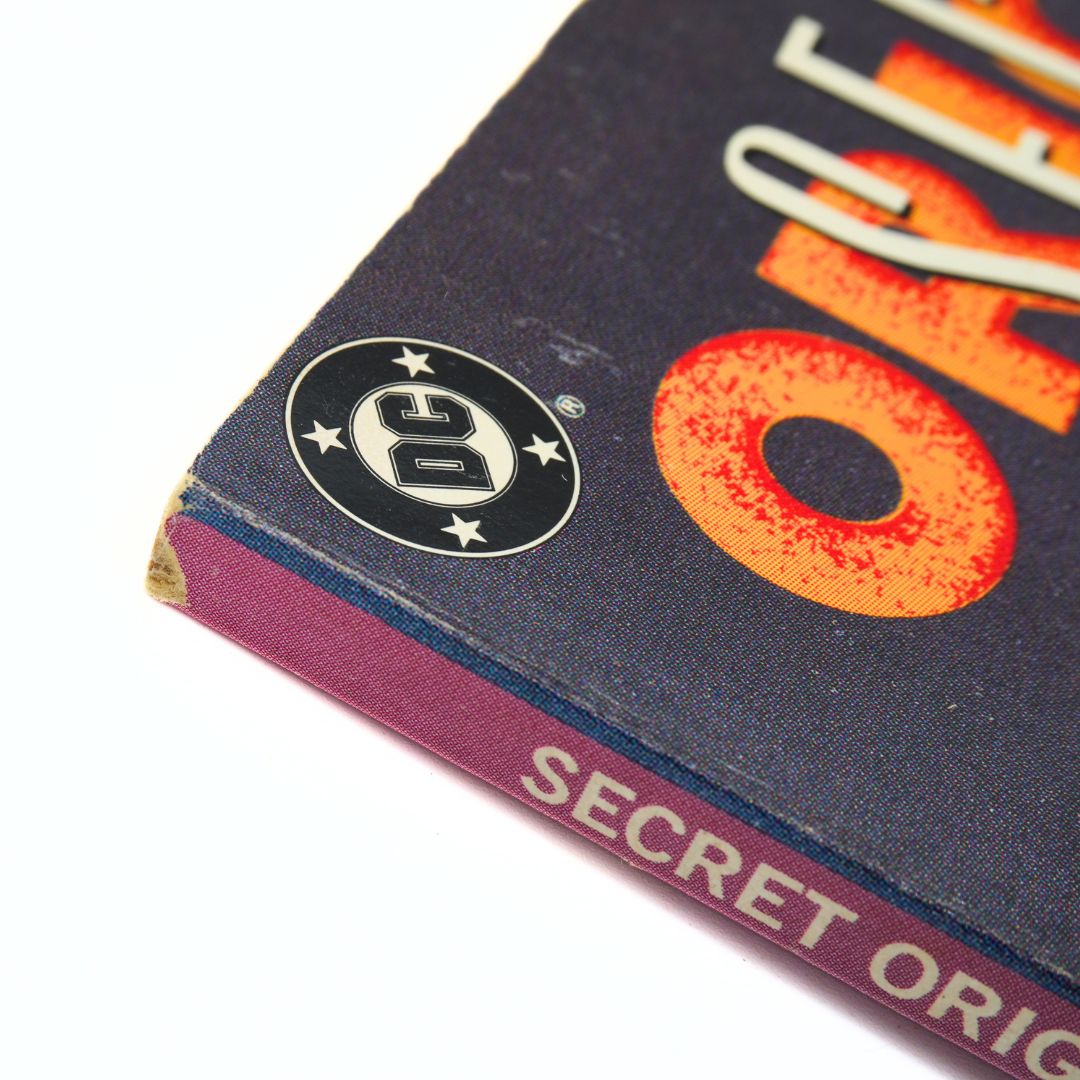 A close-up of a vintage DC Comics book, showing the dark blue cover with a prominent orange 'ONE' and a circular black-and-white DC logo sticker. The spine is visible, showing the text 'SECRET ORIG'.