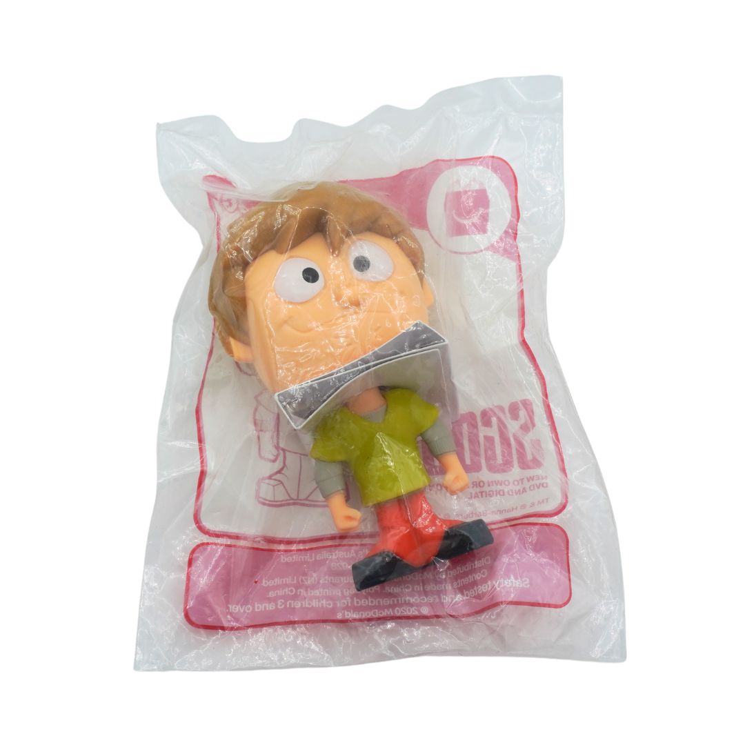 A packaged toy figurine of a cartoon boy with brown hair, wearing a green shirt and red pants, sealed inside a clear plastic bag with pink printed graphics.
