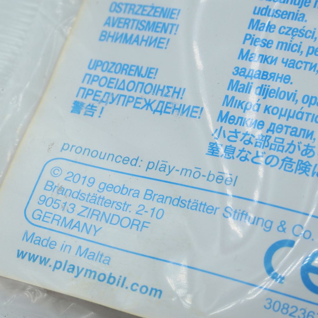 Close-up of a white plastic package with blue printed text in multiple languages, including a CE mark and the brand name geobra Brandstätter Stiftung & Co. GmbH, with Playmobil website visible.