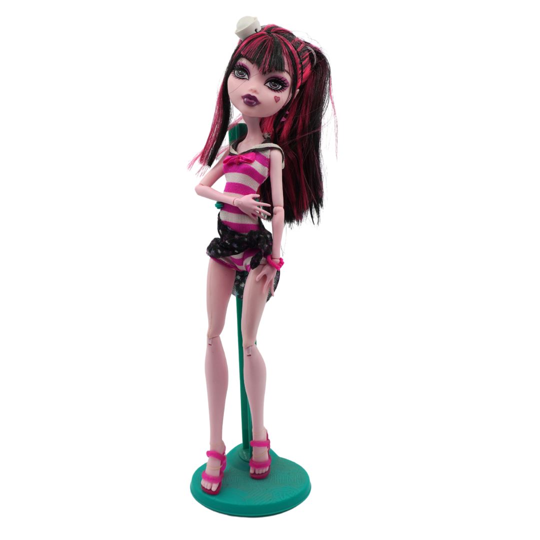 A Monster High doll with dark pink and red hair, wearing a pink and white striped top, black polka-dot shorts, and pink heels, standing on a teal base against a white background.