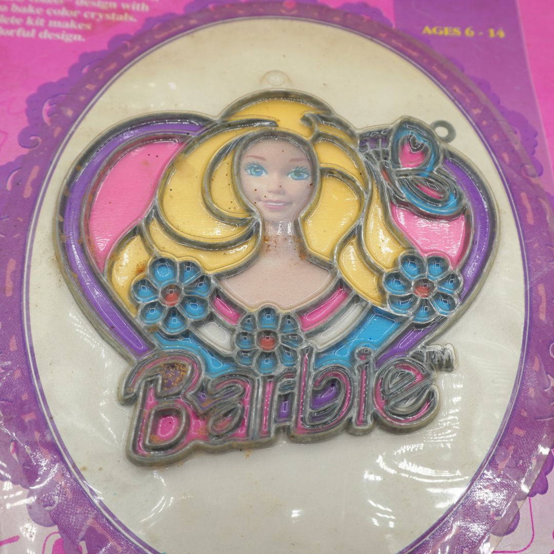 A vintage Barbie-themed stained glass ornament in its original pink and purple blister packaging. The ornament features a stylized blonde Barbie face with blue eyes, yellow hair, pink and purple heart-shaped outlines, blue flowers, and the word 'Barbie' at the bottom.