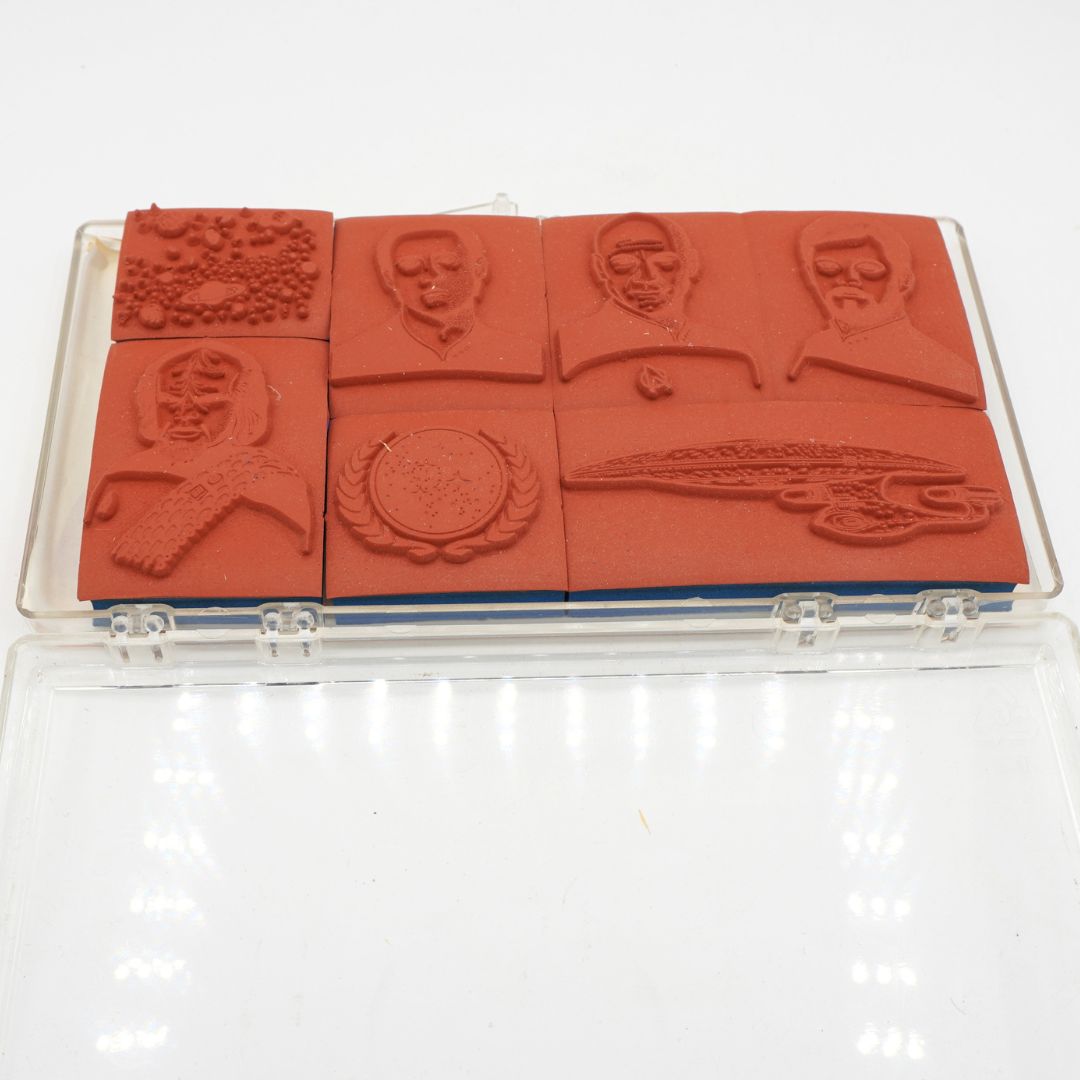 A set of six red rubber stamps in a clear plastic case, featuring portraits of historical figures, a bird, a circular emblem, and a ship design.