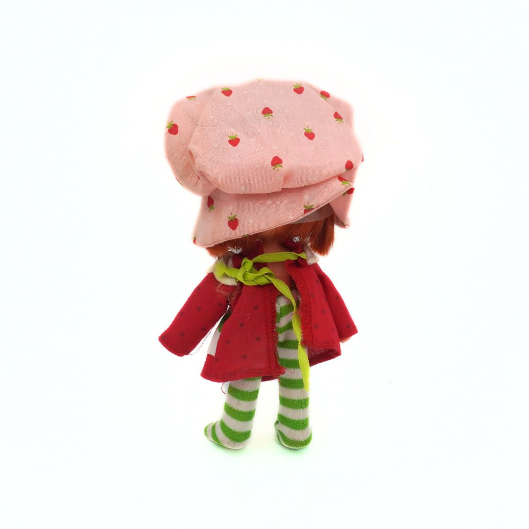 Plastic doll with a pink polka dot hat, red coat, and green striped pants on a white background