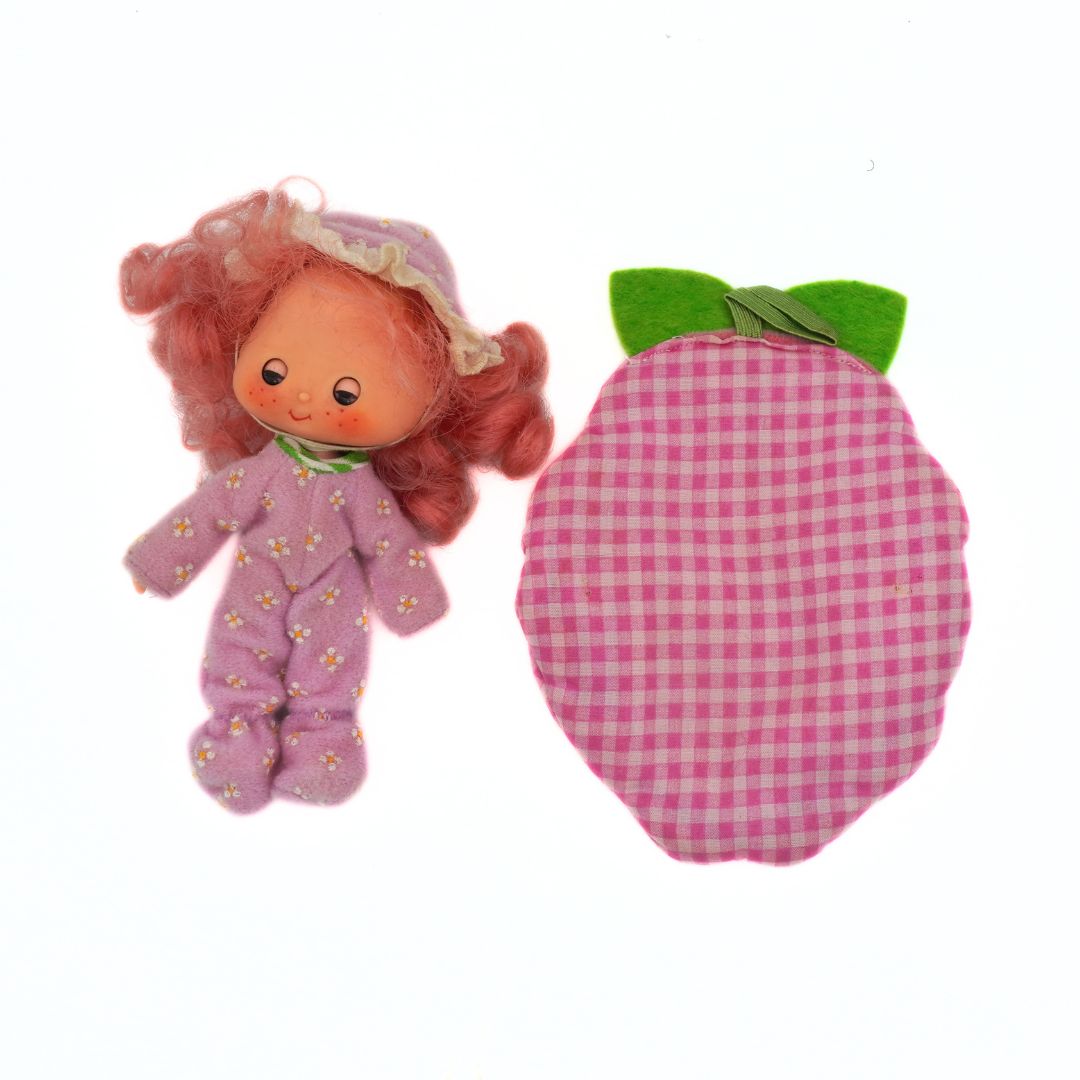 Small doll with red hair and a matching pink checkered bag on a white background