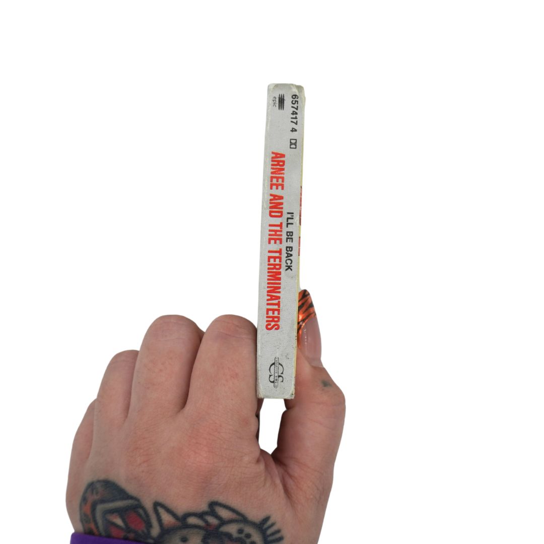 A hand holds a slim cassette against a plain white background. The spine is off-white with red text reading 'ARNEE  AND THE TERMINATORS'